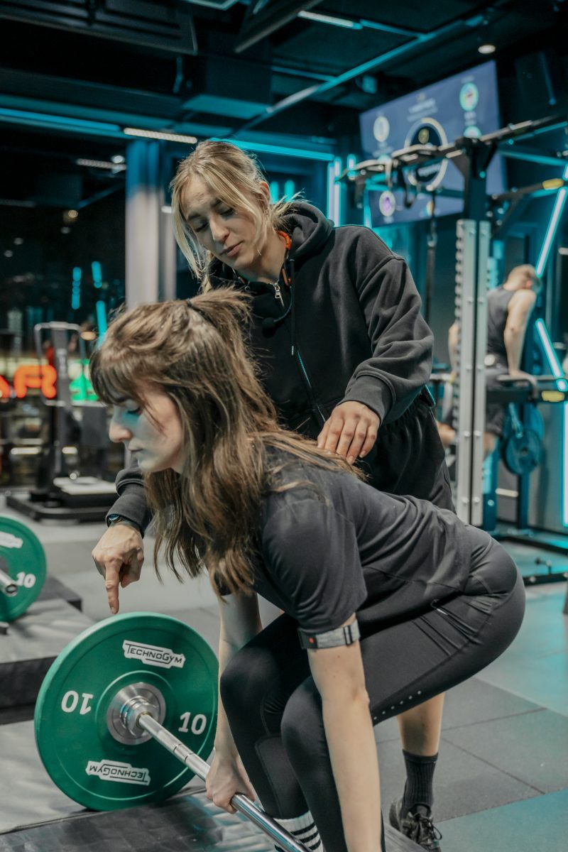 Eine junge Frau macht Deadlifts und eine Trainerin unterstützt sie dabei.